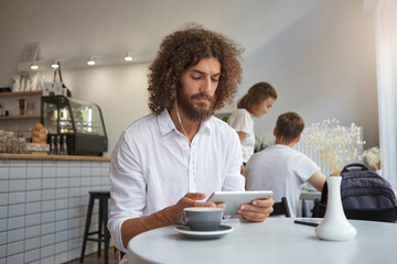 Handsome young curly man with beard sitting at table in cafe with tablet in hands, looking seriously on screen and wearing earphones, posing over city cafe background