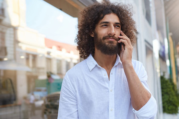 Outdoor portrait of good looking young curly male with lush beard, talking on mobile phone while having lunch break, wearing white shirt