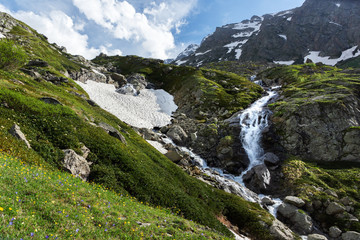 waterfall and glacier in the mountains in summer, flowering meadow, contrast between snow and green grass