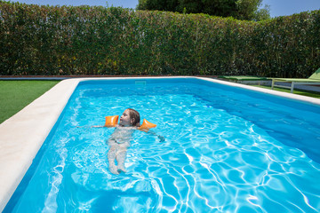relaxed four years old blonde child with orange floater sleeves in arms, armbands, swimming and floating in blue transparent water of pool