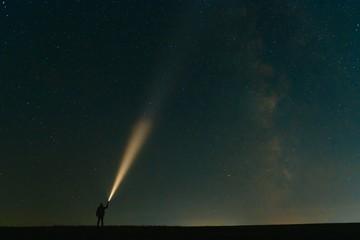Back view of man with flashlight standing on green grassy field under beautiful dark blue summer starry sky. Night photography, beauty of nature concept. Wide panorama, copy space background. - obrazy, fototapety, plakaty