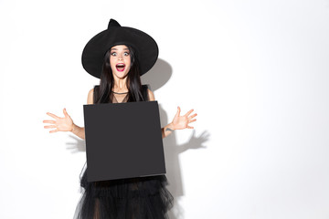 Young brunette woman in black hat and costume on white background. Attractive caucasian female model. Halloween, black friday, cyber monday, sales, autumn concept. Holding empty sheet for your ad.