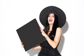 Young brunette woman in black hat and costume on white background. Attractive caucasian female model. Halloween, black friday, cyber monday, sales, autumn concept. Holding empty sheet for your ad.