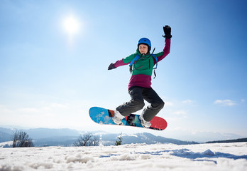 Young female snowboarder jumping in air on copy space background of clear bright blue sky and snowy...