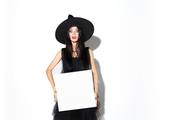 Young brunette woman in black hat and costume on white background. Attractive caucasian female model. Halloween, black friday, cyber monday, sales, autumn concept. Holding empty sheet for your ad.