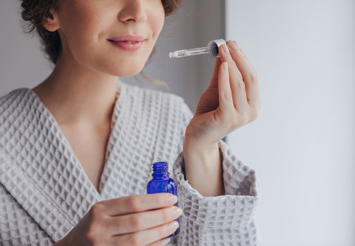 Hands of unrecognisable woman in bathrobe holding bottle of cosmetic serum and a pipette.