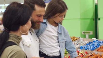 A beautiful young mother with a bearded husband and a cute seven-year-old daughter buy chocolate candies and confectionery in a large supermarket. Slow motion.