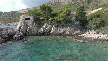 Flying over a cape on which is seen a lonely house surrounded by coniferous trees. The turquoise sea washes the rocky coast of the cape and the sandy coastline, sea waves roll on the shore