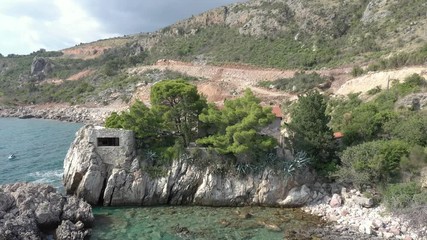 Flying over a cape on which is seen a lonely house surrounded by coniferous trees. The turquoise sea washes the rocky coast of the cape and the sandy coastline, sea waves roll on the shore