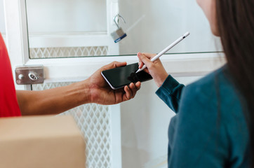 woman customer using pen signature on digital mobile phone with delivery service man in red uniform for receiving parcel post box from courier at home, express delivery and shopping online concept