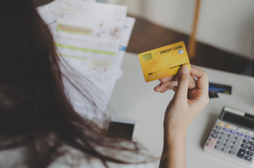 back view of young woman holding credit card and analysis with family budget cost bills and calculator on desk in home office, plan money cost saving, investment, business finance and expenses concept