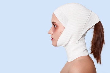 Profile of female wearing white medical bandage, looking straight ahead, posing isolated over blue studio background. Woman with pony tail having anty aging procedures. Copy space for advertisment.