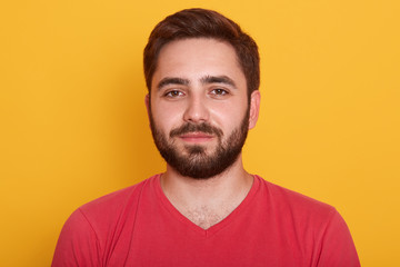 Fototapeta premium Portrait of happy man standing and looking directly at camera with pleased facial expression, bearded male wearing red casualt shirt, being in good mood, posing isolated over yellow background.