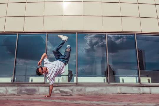 strong sporty man dancing break dance, stands one arm red-haired, young guy, free space for a text of motivation, summer city background of glass windows of building, jeans, sunglasses, white T-shirt
