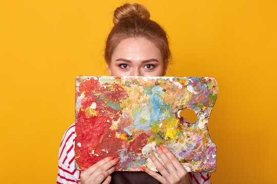Studio shot of attractive young female artist holding and hiding behind color palette or her artwork, looking at camera, adorable female wearing striped shirt, posing isolated over yellow background.