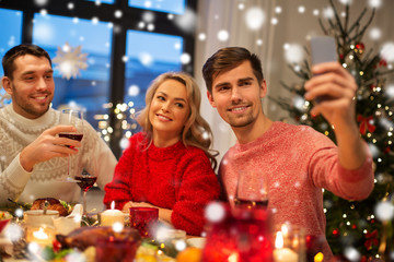 holidays and celebration concept - happy friends taking selfie by smartphone at home christmas dinner over snow