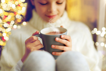 hot drinks, holidays and people concept - close up of beautiful girl in winter sweater with hot chocolate in mug sitting over christmas lights background