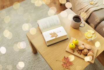 hygge and cozy home concept - book, autumn leaves, cup of tea with lemon, almond nuts and oatmeal cookies on table