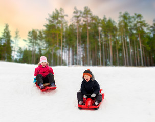 Fototapeta na wymiar childhood, sledging and season concept - happy little kids sliding on sleds down snow hill in winter over forest background