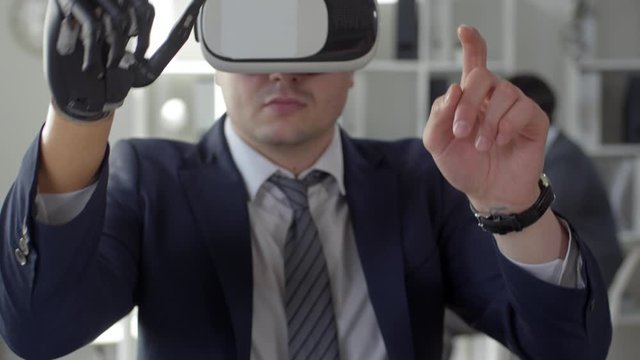 Tilt up shot of businessman with bionic prosthetic hand using VR headset, swiping and tapping on invisible touchscreen while working in the office