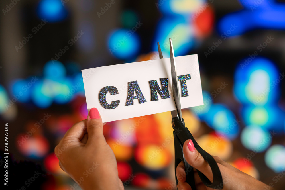 Wall mural Woman hand are cutting over white paper with scissors over the word cant and converting it to the word can. City lights bokeh as background. Motivational and inspiraton concept.