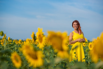 Naklejka na ściany i meble A red-haired woman in a yellow dress is standing in a field of sunflowers. Beautiful girl in a skirt sun enjoys a cloudless day in the countryside. Pink locks of hair.