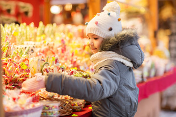 holidays, childhood and people concept - happy little girl choosing sweets at christmas market in...