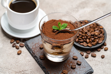 Portion of Classic tiramisu dessert in a glass cup on stone serving board and fres espresso coffee on concrete background