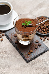 Portion of Classic tiramisu dessert in a glass cup on stone serving board and fres espresso coffee on concrete background