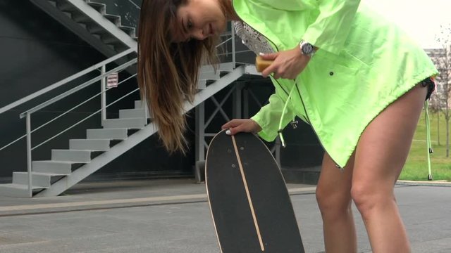 Beautiful girl in a green jacket rubs a longboard. She's getting ready to ride on a skateboard.