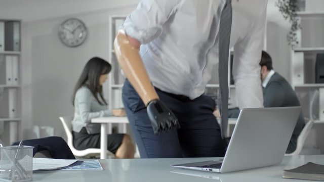 Caucasian businessman walking to his workplace, taking off prosthetic bionic hand, putting it on office desk and typing on laptop