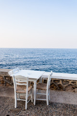Weiße leere Stühle und Tisch aus Holz stehen an einer Strandpromenade mit Ausblick aufs Meer