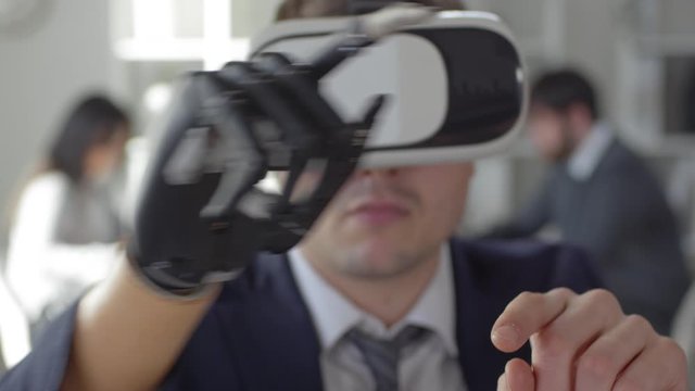 Caucasian businessman with bionic prosthetic hand using VR headset, swiping and tapping on invisible touchscreen at office desk