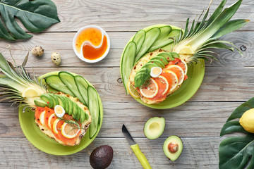 Top view on pineapple boats with smoked salmon and avocado slices with lemon and quail eggs, flat lay on old wooden table