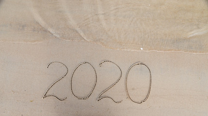 Handwritten inscription 2020 on beautiful sand beach. Lettering on beach with wave and clear blue sea. New Years 2020 concept.