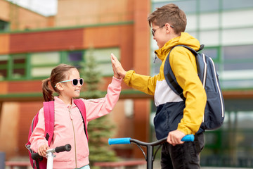 education, childhood and people concept - happy school children with backpacks and scooters making high five outdoors