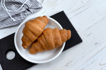 Two croissants on white plate on black cutting board and white wooden table Top view 
