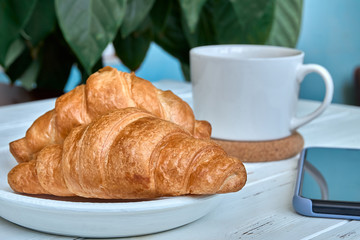 Croissants on white plate, a cup of coffee and smartphone on white wooden table.  Continental European breakfast concept