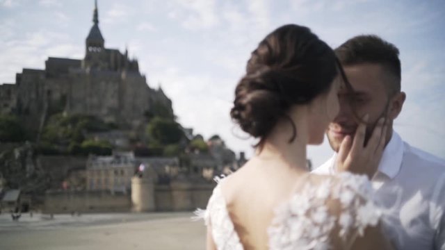 Close-up view of beautiful romantic wedding couple standing with famous Mont Saint-Michel abbey on the background in summer day against blue sky. Action. Sensual wedding day