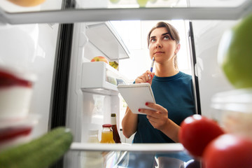 healthy eating and diet concept - woman opening fridge and making list of necessary food at home...