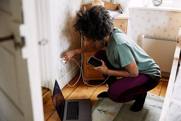 Side view of young woman plugging mobile phone charger in electrical outlet at home seen through doorway