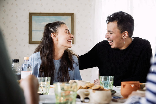 Cheerful friends looking at each other while sitting by food and drink in house