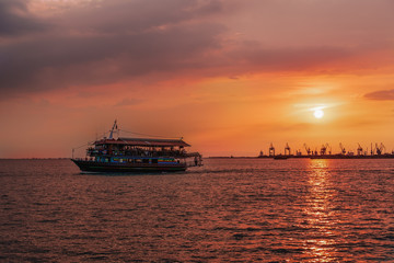 Tourist ship sailing during a golden hour sunset at Thessaloniki, Greece. Unidentified crowd aboard a small ship at Thermaikos gulf during orange sunset, with port cranes background.