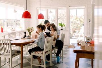 Father looking at girl using digital tablet while sitting with daughter doing homework at dining table