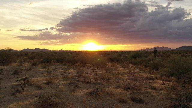 A drone moving back away from a saguaro cactus and then off to the right as the cactus passes in front of a sunset with clouds and mountains of the Sonoran Desert in the distance.