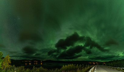 Strong bright Aurora Borealis behind heavy clouds over road, Joesjo Lake and Scandinavian mountains in Swedish Lapland Lake look very dramatic, Northern Sweden. Late summer night.