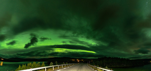 Panoramic photo of strong bright Aurora Borealis behind heavy clouds over road, Joesjo Lake and Scandinavian mountains in Swedish Lapland look very dramatic, Northern Sweden. Late summer night.