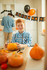 Portrait of cheerful boy sitting at the table with pumkins and preparing for Halloween party with fis father standing in the background