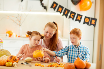 Young smiling mother helping to her daughter and son to make Jack O' Lantern from pumpkins while they sitting at the table at home