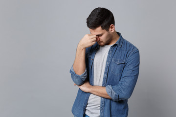 Handsome tired stylish unshaven young man in denim jeans shirt posing isolated on grey wall background studio portrait. People lifestyle concept. Mock up copy space. keep eyes closed, put hand on nose